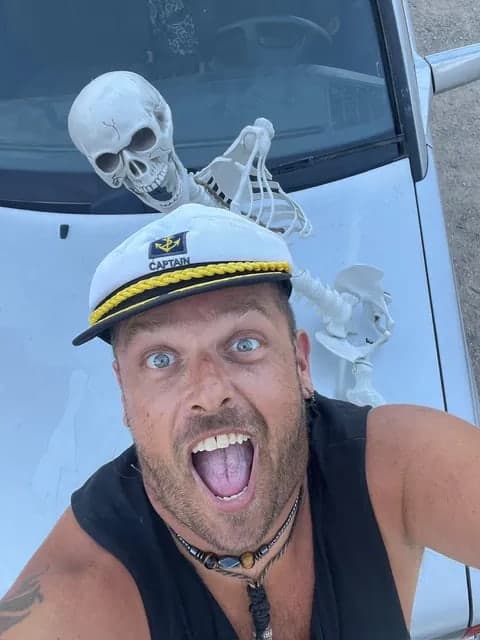 CaptainGee in a white CAPTAIN hat with his mouth wide open, a skeleton prop lying on the hearse bonnet behind him
