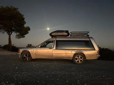 Silver Mercedes hearse camper with a roof box, roof basket and alloy wheels, parked on gravel at dusk with the moon above