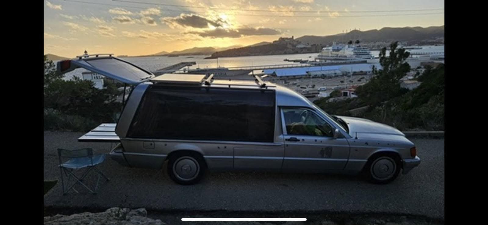 Silver Mercedes hearse camper with rear hatch open and slide-out platform, camp chair, coastal Ibiza overlook at golden hour