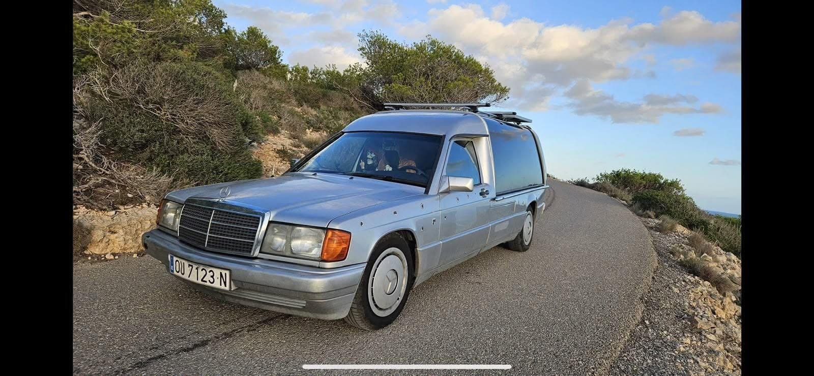Silver Mercedes hearse camper with bullet-hole decals and skeleton co-pilot, parked on a winding Spanish coastal road